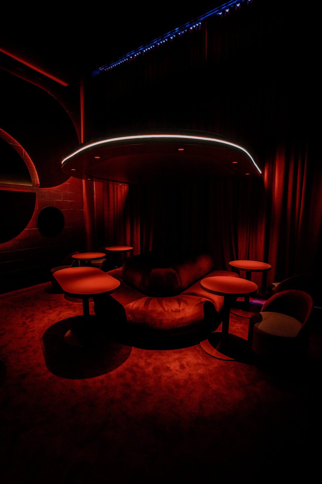 A low-lit corner of Alter Ego with round red tables and a curved sofa beneath a glowing ceiling light, framed by draped curtains